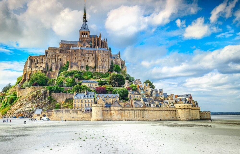 Mont Saint-Michel, abbazia e villaggio storico della Normandia, si erge in cima a un'isola rocciosa circondata da pianure sabbiose e acqua sotto un cielo blu vibrante: un'attrazione da non perdere in qualsiasi tour di Capodanno nella regione.
