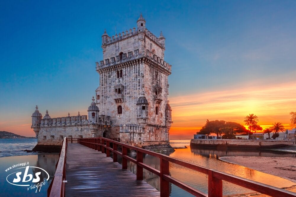La Torre di Belém a Lisbona, Portogallo, si erge sul fiume al tramonto, con una passerella di legno che conduce alla storica fortezza di pietra. Le palme e i colori vivaci riempiono il cielo: una tappa obbligata di ogni Tour Eleganza.