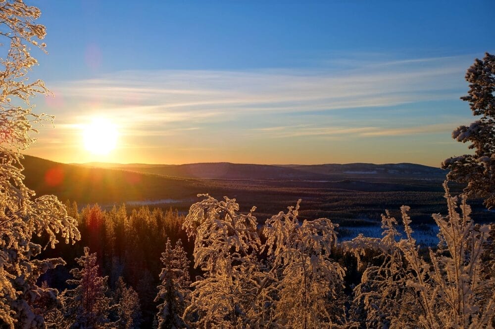 Il sole tramonta su un paesaggio invernale, illuminando gli alberi innevati e proiettando una calda luce sulla foresta e sulle colline lontane sotto un cielo azzurro e limpido: una scena perfetta per un momento di Bozza automatica.