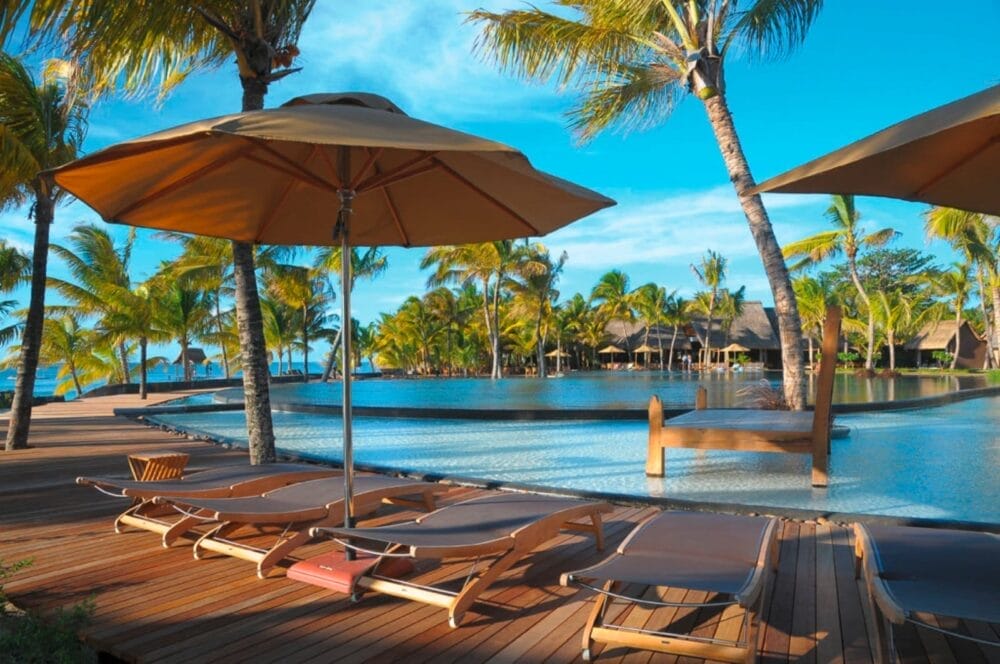 Sedie a sdraio e ombrelloni si trovano su un ponte di legno accanto alla piscina scintillante del Beachcomber Resort Trou Aux Biches, circondato da palme e capanne tropicali con l'oceano sullo sfondo sotto un cielo blu brillante.