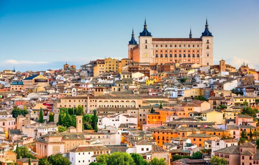 Una vista panoramica di Toledo, in Spagna, con edifici storici dalle facciate colorate e l'imponente fortezza dell'Alcázar che sovrasta la città, con Madrid a breve distanza sotto un cielo azzurro e limpido.