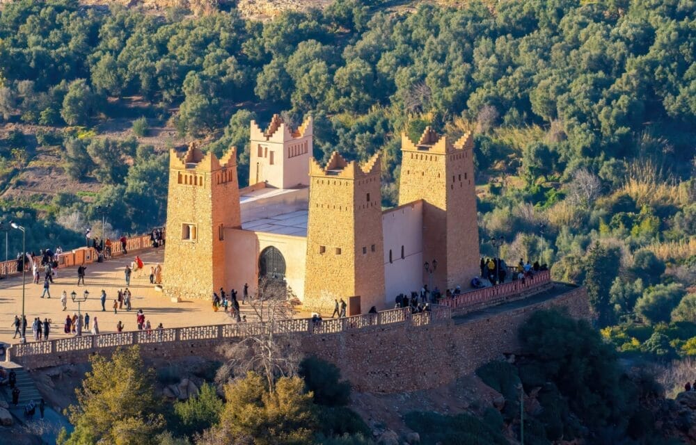 Un edificio rettangolare, simile a una fortezza, con quattro alte torri quadrate, si trova su una collina circondata da alberi. Molte persone passeggiano sull'ampia terrazza, assaporando la vista panoramica e ammirando il design unico della Bozza automatica.