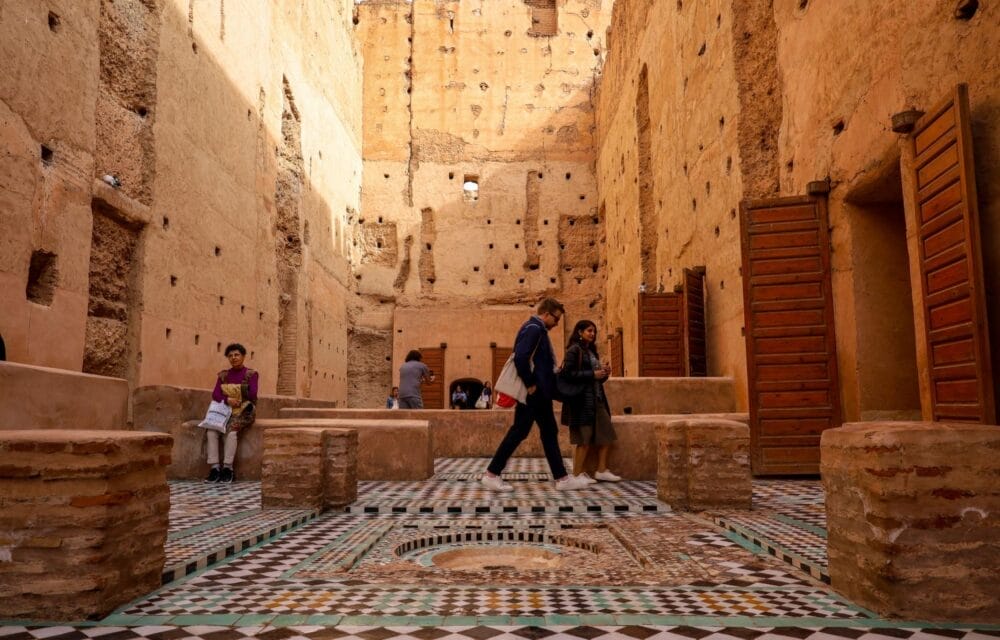 I visitatori esplorano le rovine di un edificio storico con pareti alte e strutturate e pavimenti di piastrelle a mosaico colorate. La Bozza automatica si nota vicino alle porte di legno aperte e alle colonne di mattoni, mentre alcune persone sono sedute o camminano all'interno.