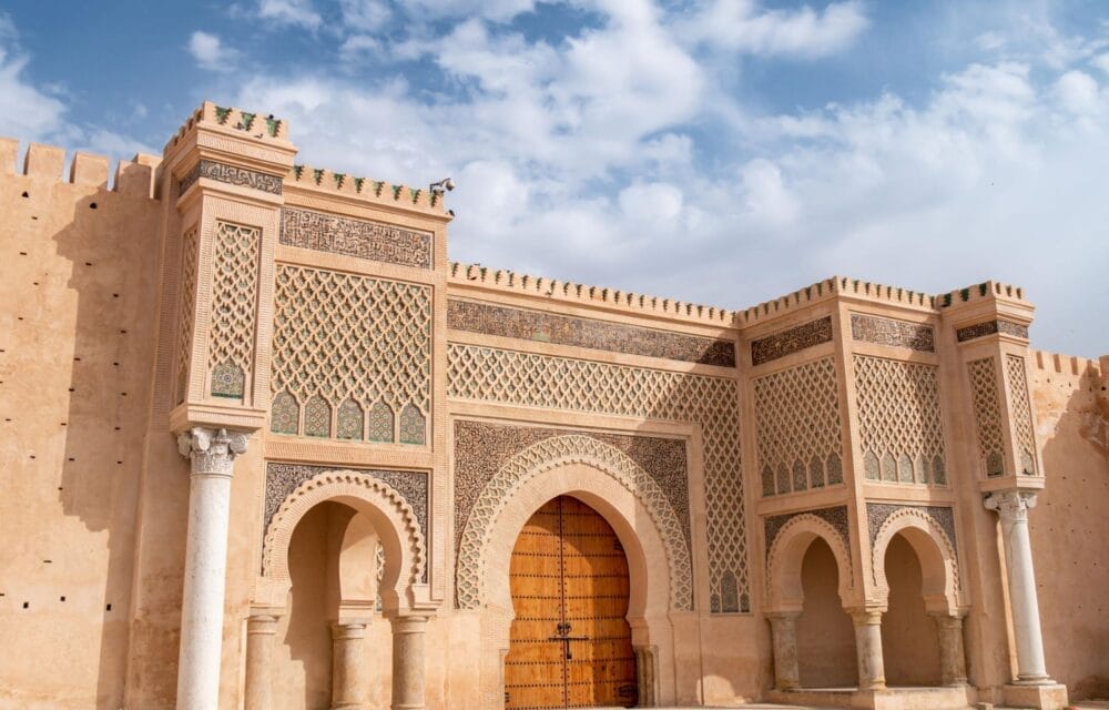 Una porta storica e ornata della città marocchina, con Bozza automatica, motivi geometrici dettagliati, porte ad arco, intricate piastrelle e colonne decorative, sullo sfondo di un cielo parzialmente nuvoloso.