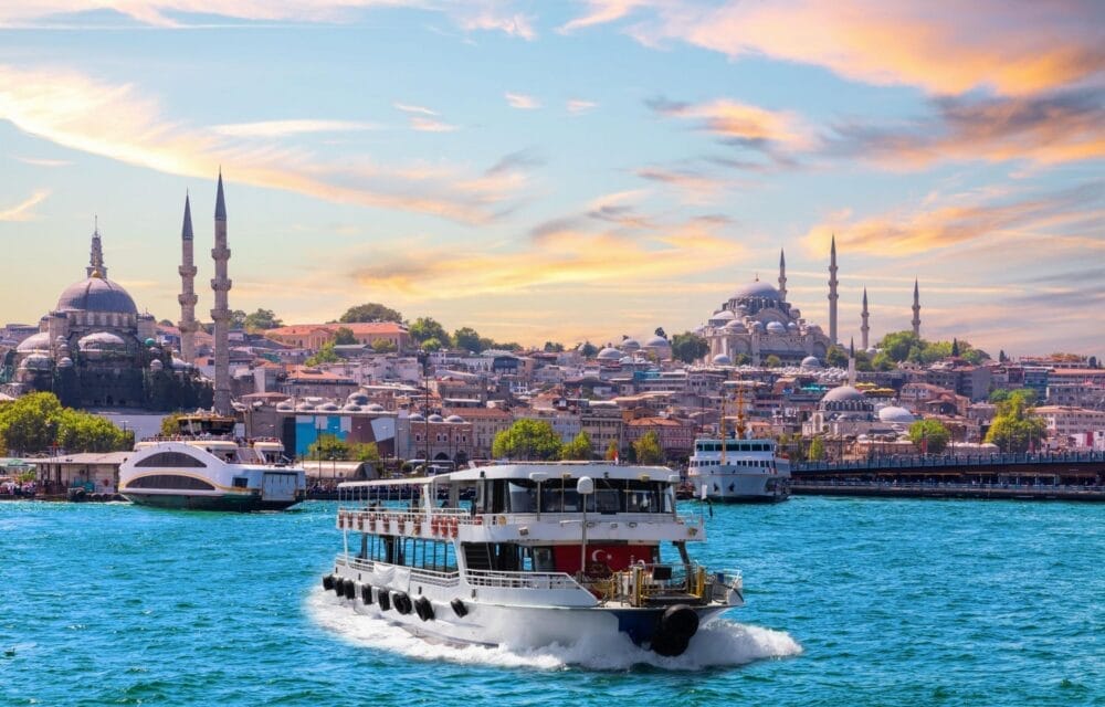 Un traghetto passeggeri naviga sull'acqua blu brillante con il paesaggio urbano di Istanbul e le moschee storiche sullo sfondo, sotto una bozza automatica di cielo colorato e parzialmente nuvoloso al tramonto.