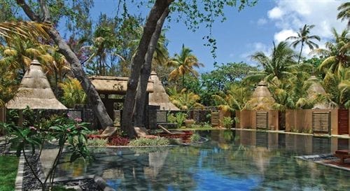 Una serena fuga tropicale, lo Shandrani Beachcomber è caratterizzato da edifici con tetti di paglia, palme lussureggianti e una piscina riflettente sotto un cielo blu brillante con nuvole sparse.