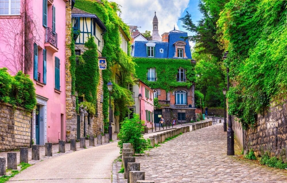 Una strada acciottolata di Parigi, fiancheggiata da edifici colorati, tra cui facciate rosa e ricoperte di edera, curva dolcemente verso l'alto. Il verde lussureggiante e gli alberi circondano l'area, con una torre lontana visibile sotto un cielo parzialmente nuvoloso.