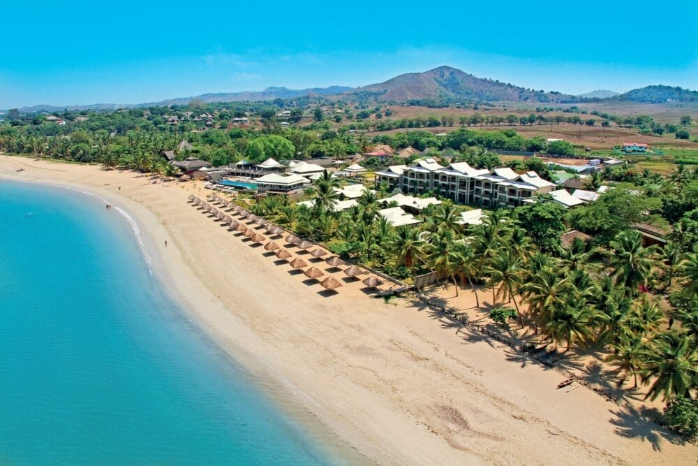 Veduta aerea del Veraclub Palm Beach, con acqua cristallina, file di ombrelloni di paglia, palme ondeggianti e un complesso di resort immerso in una vegetazione lussureggiante con colline lontane sotto un cielo azzurro brillante.