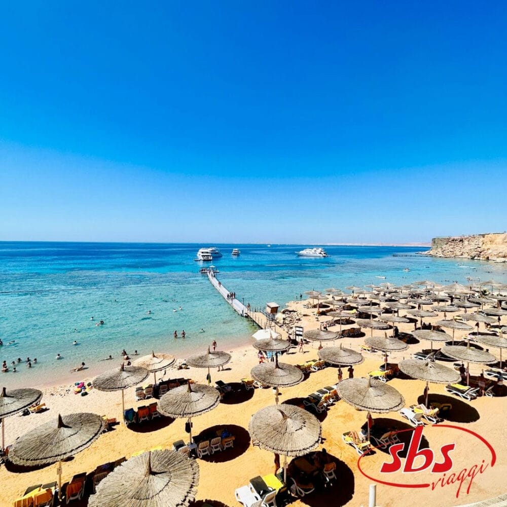 Una spiaggia soleggiata di Sharm el Sheikh con ombrelloni di paglia e sedie a sdraio, persone che nuotano nell'acqua limpida e blu e barche ancorate vicino a un molo di legno. Il logo "sbs viaggi" è in basso a destra. 2023.