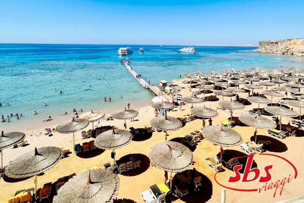 Una spiaggia soleggiata di Sharm el Sheikh con ombrelloni di paglia, lettini e persone che fanno il bagno. Un molo di legno si estende nell'acqua limpida e azzurra, con barche ancorate nelle vicinanze. Il logo di SBS Viaggi e il 2023 appaiono nell'angolo in basso a destra.