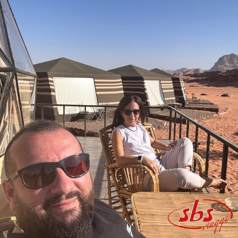 Un uomo e una donna con gli occhiali da sole si rilassano su una terrazza di legno con sedie di vimini. Dietro di loro ci sono tende a strisce in un paesaggio desertico sotto un cielo azzurro, perfetto per il vostro Tour in Giordania 2023. Il logo "sbs" è visibile nell'angolo.