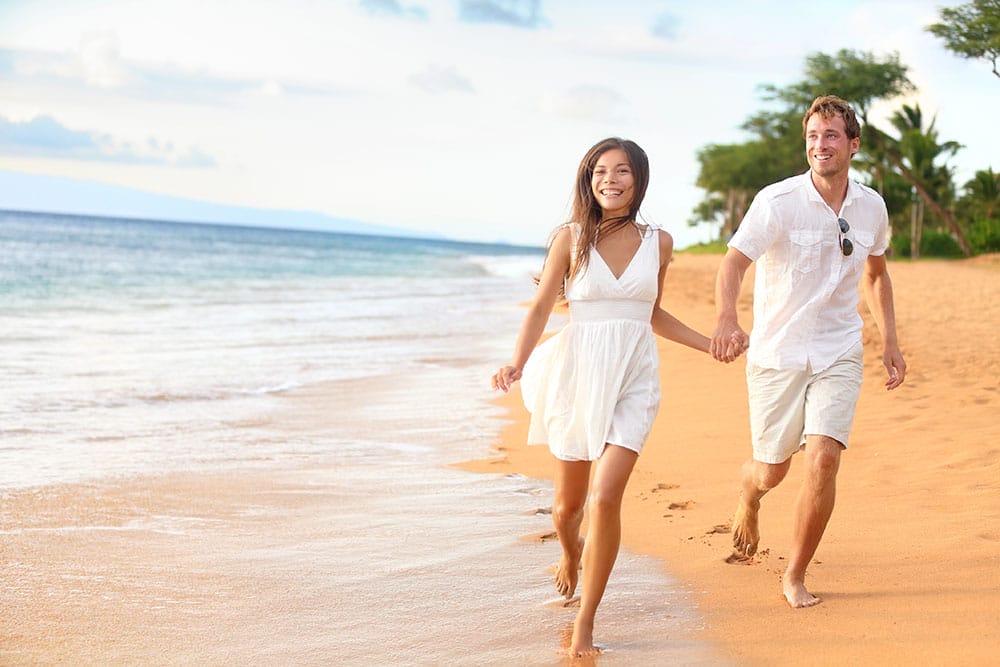 Una coppia sorridente in abiti estivi bianchi si tiene per mano e cammina a piedi nudi lungo una spiaggia sabbiosa in riva all'oceano, godendosi una Luna di Miele Su Misura, con alberi e cielo blu sullo sfondo.