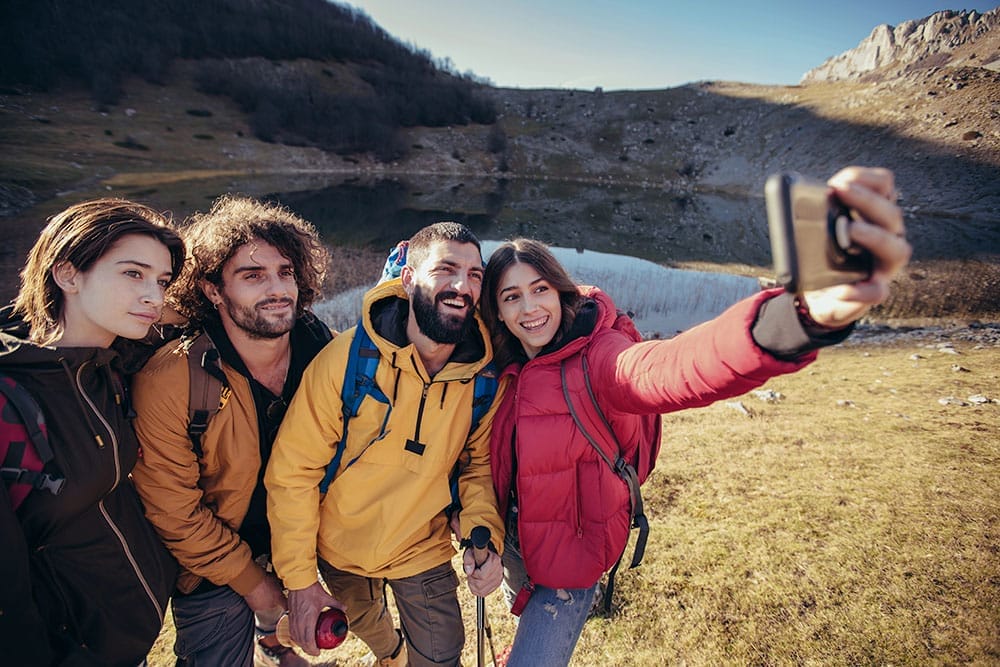 Quattro amici in tenuta da trekking stanno vicini vicino a un lago e alle montagne, sorridendo mentre uno scatta un selfie con lo smartphone. Questo momento gioioso cattura lo spirito dei viaggi per single sotto un cielo limpido e soleggiato.