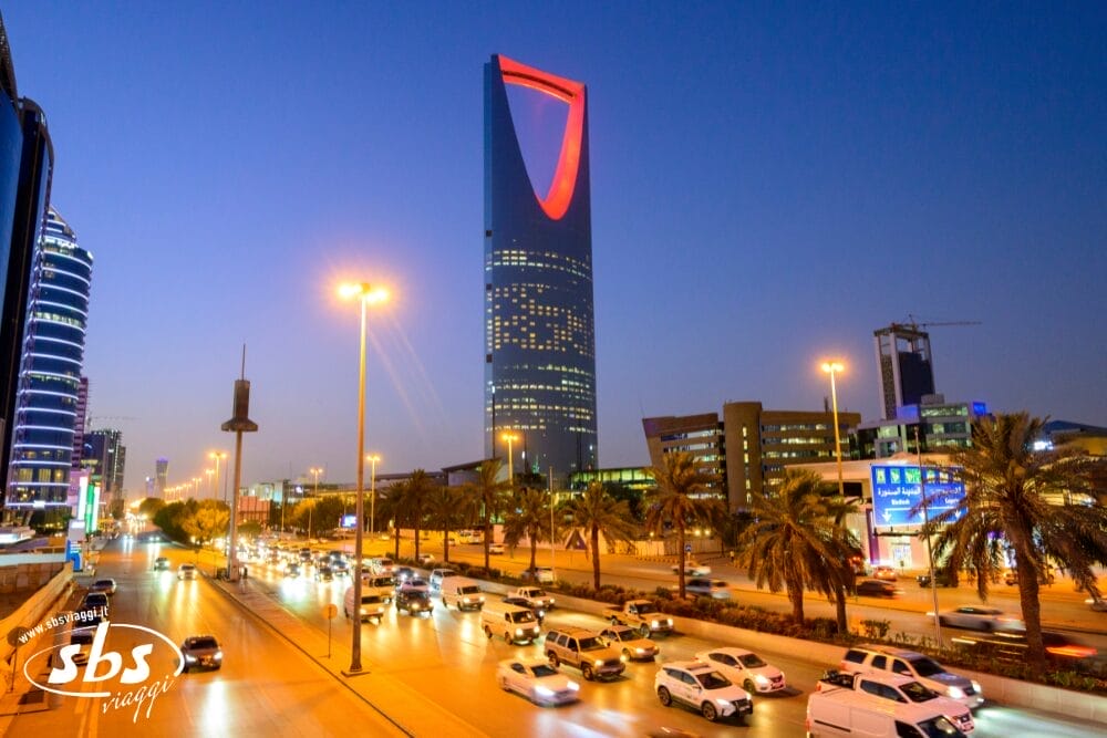 Una strada trafficata di Riyadh al tramonto, con auto e palme lungo la strada. La Kingdom Centre Tower illuminata si staglia nel cielo serale, circondata da edifici moderni e lampioni: una bozza urbana automatica.