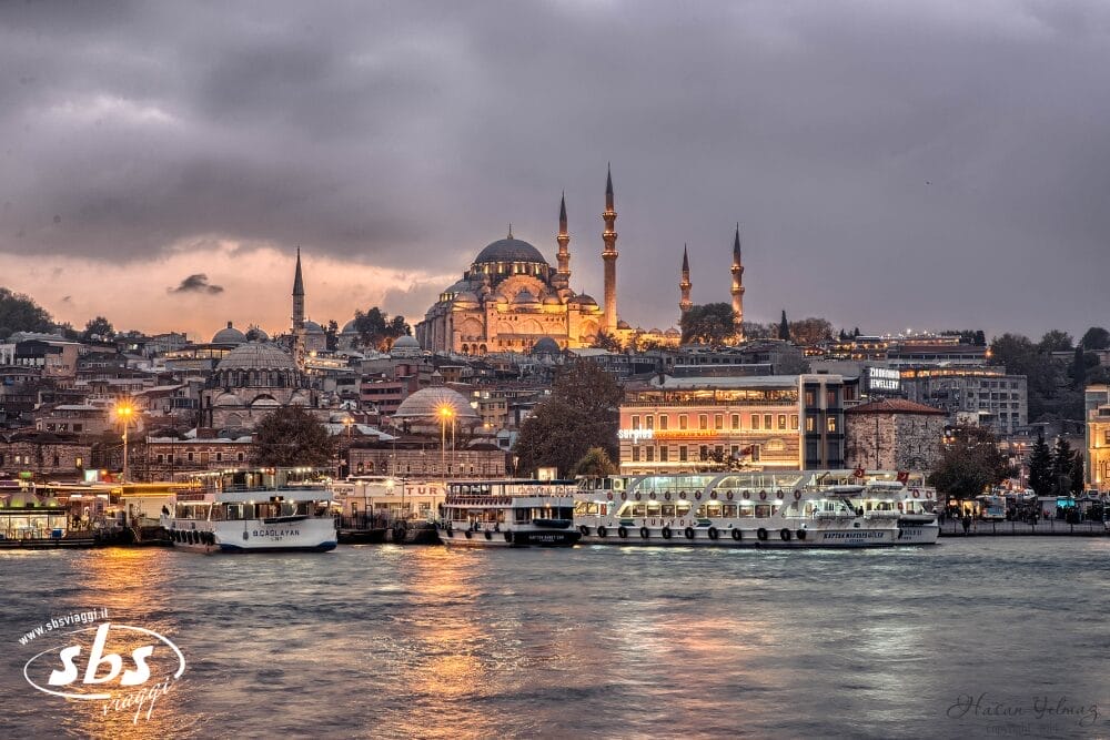La Moschea di Suleymaniye, illuminata, domina lo skyline di Istanbul al tramonto, con le barche attraccate sul lungomare e gli edifici della città in primo piano sotto un cielo nuvoloso, creando una scena che ricorda una bozza automatica.