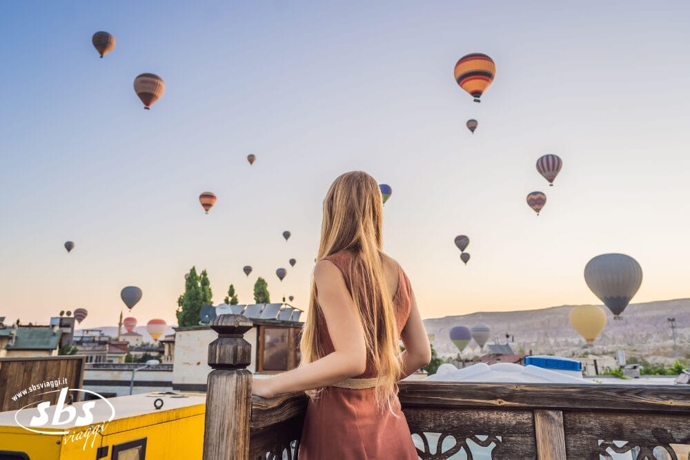 Una donna con i capelli lunghi si trova su una terrazza panoramica, ammirando la bozza automatica di numerose mongolfiere che fluttuano nel cielo durante l'alba o il tramonto su un paesaggio panoramico.