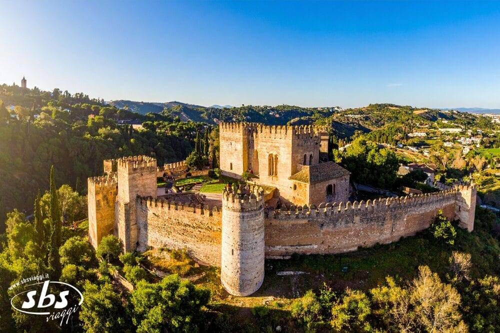 Veduta aerea di un grande castello medievale in pietra a Malaga, con alte torri e mura merlate, circondato dal verde e da dolci colline sotto un cielo azzurro e limpido. Il logo "sbs" appare nell'angolo in basso a sinistra. Gran Tour Andalusia.