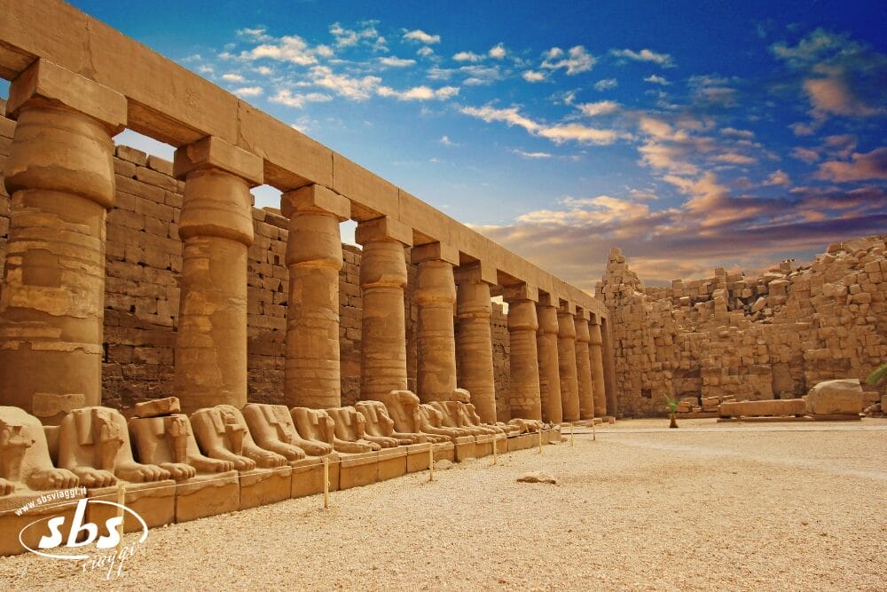 Antiche colonne di pietra e file di statue di sfingi, ormai logore, fiancheggiano un cortile del complesso del Tempio di Karnak a Luxor, in Egitto, sotto un vibrante cielo di Bozza automatico blu e arancione al tramonto.