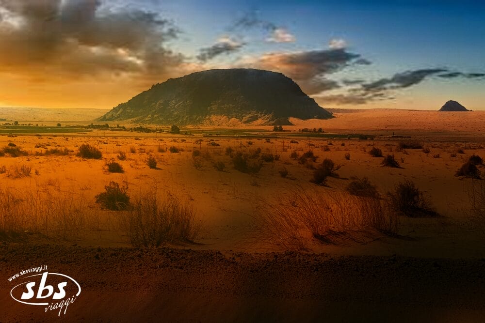 Un vasto paesaggio desertico al tramonto, con una grande collina rocciosa in lontananza, arbusti sparsi in primo piano e un cielo parzialmente nuvoloso. L'immagine è caratterizzata da una calda luce arancione e dal logo "Bozza automatica sbs viaggi" nell'angolo in basso a sinistra.