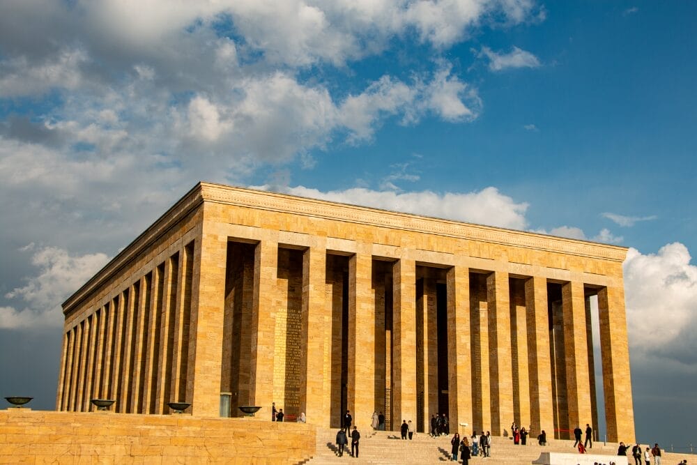 Un grande mausoleo rettangolare in pietra con alte colonne si erge sotto un cielo parzialmente nuvoloso, con persone che camminano e si raccolgono vicino ai suoi gradini, come se fossero in pausa in un momento di bozza automatica.