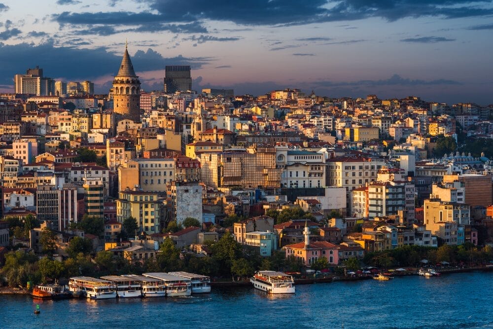 Un paesaggio urbano di Istanbul al tramonto, con la Torre di Galata che si erge al di sopra di un gruppo di edifici storici, diverse imbarcazioni attraccate sul lungomare del Corno d'Oro e lo skyline immerso in un tenue bagliore automatico di bozza.