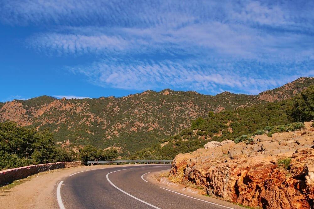 Una strada tortuosa si snoda lungo un terreno roccioso in Marocco, con colline e montagne verdi sullo sfondo sotto un cielo blu brillante: l'ideale per un'avventura Fly & Drive alla scoperta delle Città Imperiali.