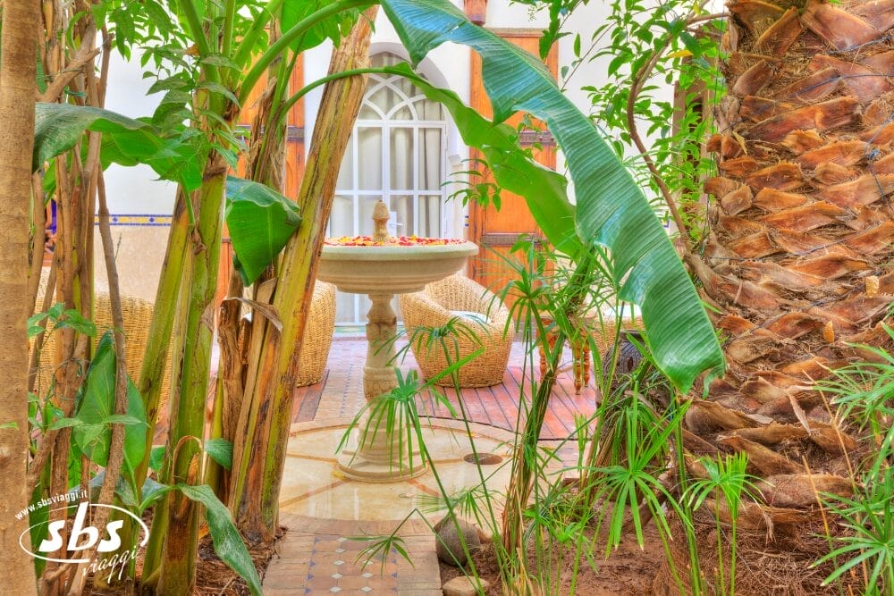 Piante verdi e palme incorniciano un cortile con una fontana in pietra al centro, circondata da sedie in vimini e da un pavimento piastrellato, creando una Bozza automatica dall'atmosfera serena e tropicale.
