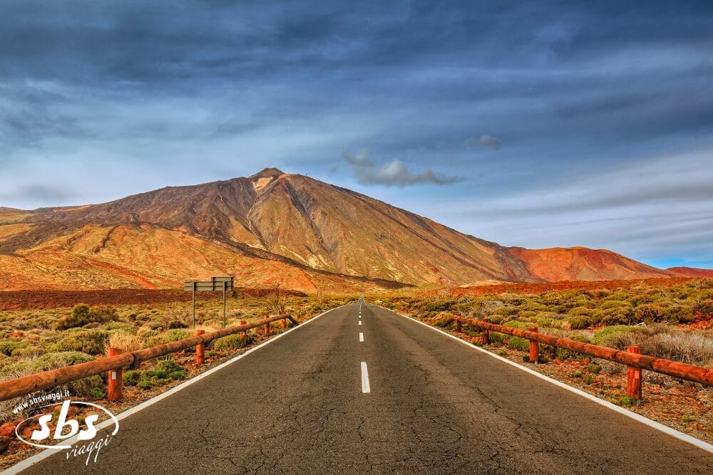 Una strada dritta conduce verso una grande montagna rocciosa sotto un cielo azzurro con nuvole sottili, circondata da una vegetazione secca e colorata e da ringhiere di legno lungo il ciglio della strada: una vista panoramica ideale per immortalare la vostra prossima Bozza automatica.