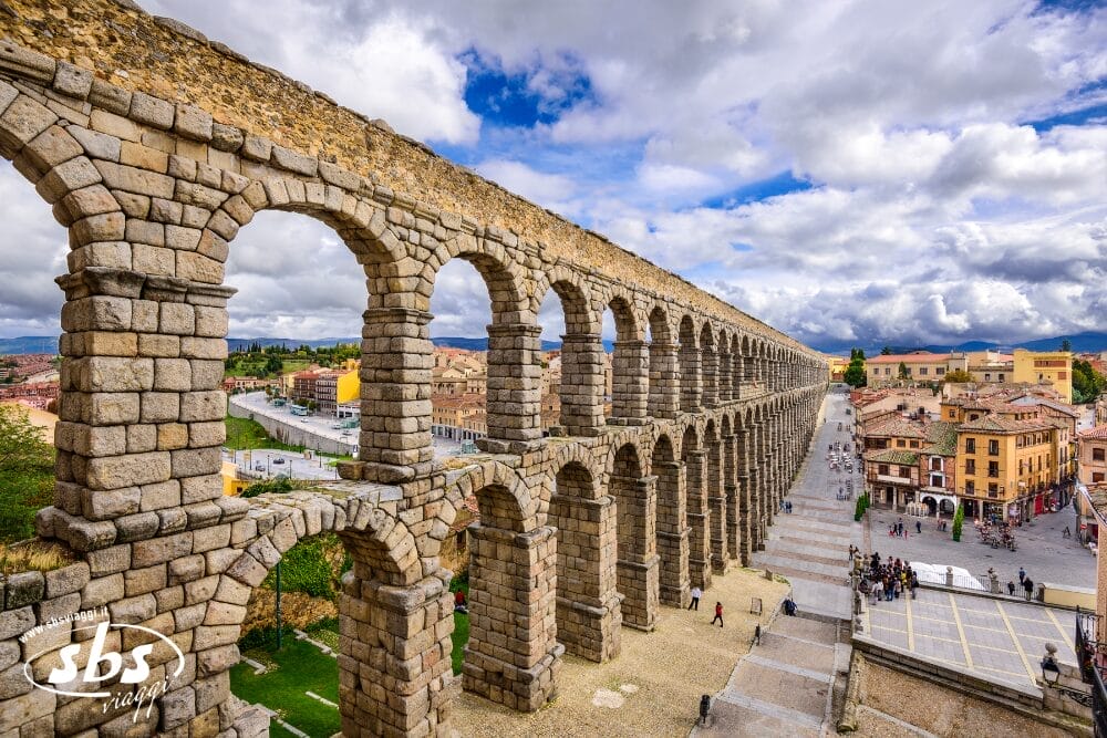 Un antico acquedotto in pietra con grandi archi si estende su un paesaggio urbano a Segovia, in Spagna, con persone che camminano al di sotto ed edifici colorati sullo sfondo sotto un cielo parzialmente nuvoloso, ripreso come bozza automatica.