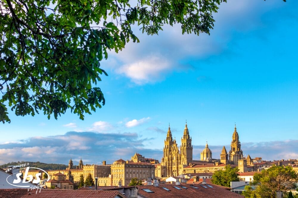 L'immagine mostra lo skyline di Santiago de Compostela, in Spagna, con le torri gemelle della storica cattedrale illuminate dalla luce del sole sotto un cielo azzurro con nuvole sparse e rami di alberi in primo piano: una scena perfetta per una Bozza automatica.