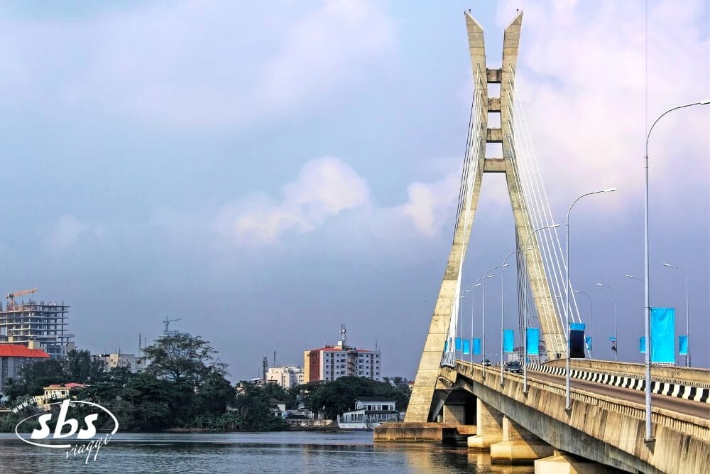 Un moderno ponte strallato attraversa l'acqua e conduce verso lo skyline di una città con alti edifici. Striscioni blu e una scritta "Bozza automatica" costeggiano il ponte, mentre il cielo è parzialmente nuvoloso. La foto include una filigrana "sbs" nell'angolo in basso a sinistra.