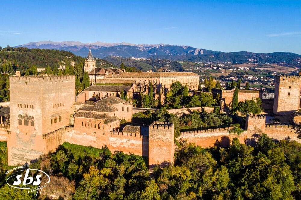 Veduta aerea dell'Alhambra, un palazzo storico e un complesso di fortezze dalle mura rossastre, circondato da alberi e montagne a Granada, in Spagna, sotto un cielo azzurro e limpido: una Bozza automatica del patrimonio davvero straordinaria.