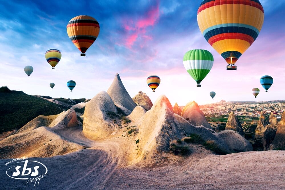 Coloratissime mongolfiere fluttuano sullo straordinario paesaggio roccioso della Cappadocia, in Turchia, al sorgere del sole. Il cielo rosa e viola crea una scena da sogno mentre il sole illumina le formazioni appuntite in questo momento magico, quasi come una bozza automatica.