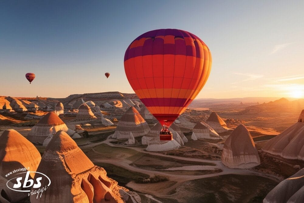 Una mongolfiera arancione e viola fluttua sopra formazioni rocciose uniche al sorgere del sole, con diverse altre mongolfiere in lontananza attraverso il paesaggio panoramico e arido. Il logo SBS Viaggi e la filigrana Bozza automatica appaiono nell'angolo in basso a sinistra.