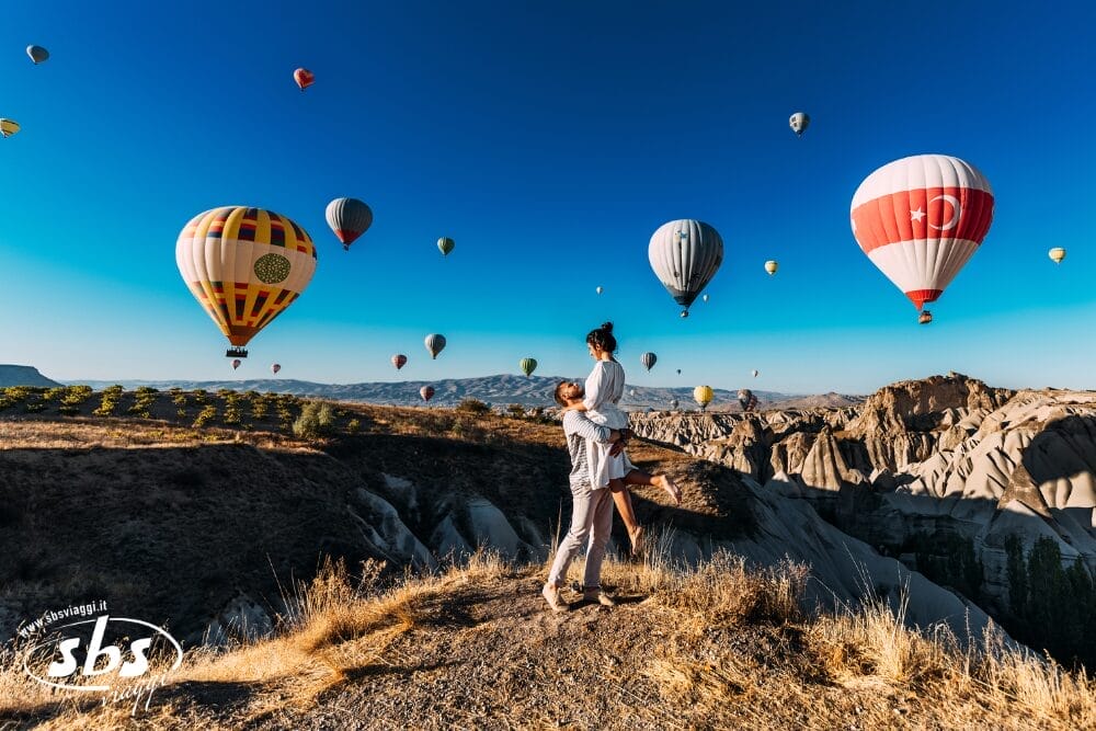 Una coppia in piedi su una collina rocciosa, con la persona che solleva il partner con gioia. Bozza automatica: mongolfiere colorate fluttuano in un cielo azzurro sopra il paesaggio roccioso sullo sfondo.
