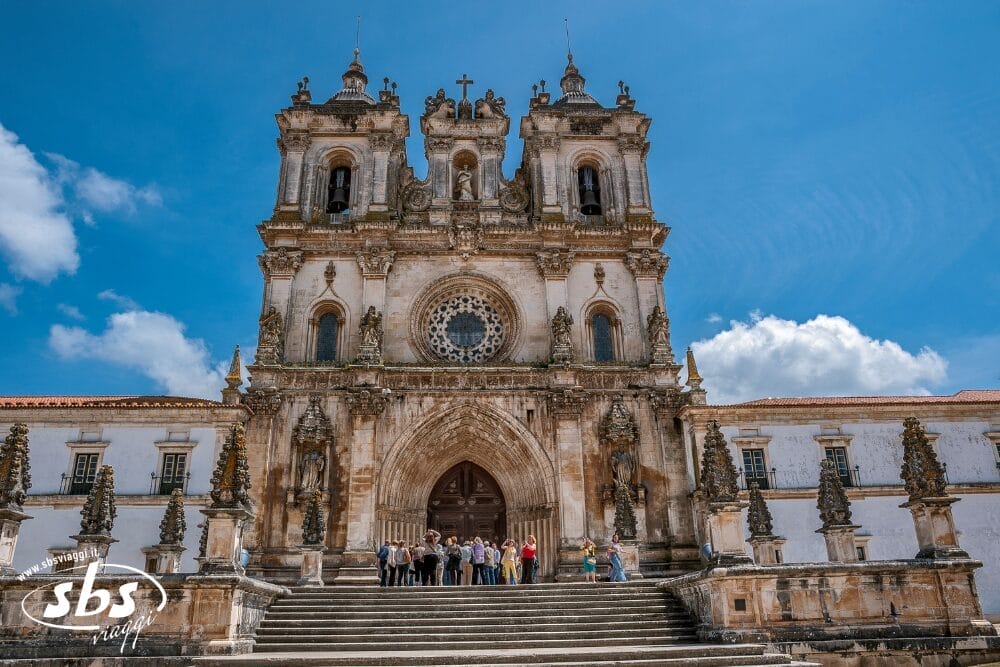 Un gruppo di persone si trova sui gradini di pietra davanti alla facciata ornata del monastero di Alcobaça, in Portogallo, con due campanili gemelli, un rosone e portali ad arco sotto un cielo azzurro: un vero momento di Bozza automatica.