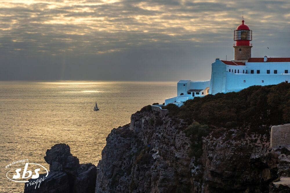 Un faro bianco con cupola rossa si erge su una scogliera rocciosa al tramonto. Il mare sottostante riflette la luce del sole, mentre una barca a vela va alla deriva nelle vicinanze: una tranquilla scena di Bozza automatica sotto un cielo parzialmente nuvoloso.