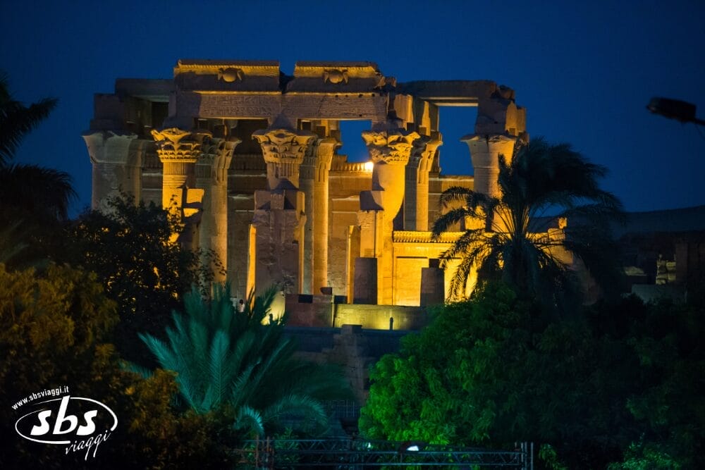 Antiche rovine di un tempio illuminate di notte da luci dorate, circondate da alberi scuri e fronde di palma, con un cielo blu intenso di Bozza automatica sullo sfondo. Il logo di un'agenzia di viaggi è visibile nell'angolo in basso a sinistra.