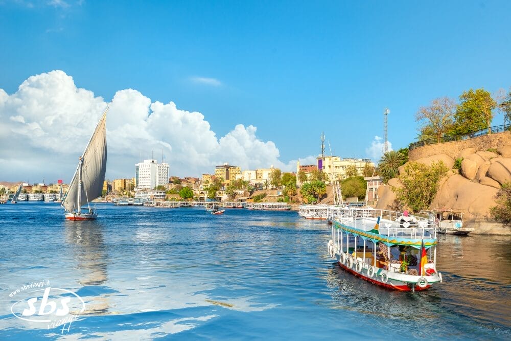 Barche colorate navigano sul fiume Nilo vicino a un paesaggio urbano con edifici bianchi e gialli, colline rocciose e alberi rigogliosi sotto un cielo blu brillante con nuvole soffici: una scena vibrante come una bozza automatica di un capolavoro della natura.