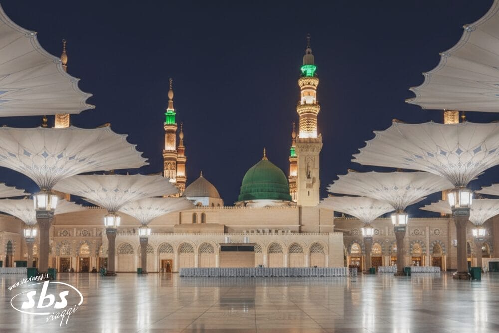 Al-Masjid an-Nabawi (Moschea del Profeta) di Medina illuminata di notte, con grandi ombrelli retrattili, colonnati ad arco, cupole e minareti verdi e illuminati d'oro. Il tranquillo cortile sembra in attesa di una bozza automatica.