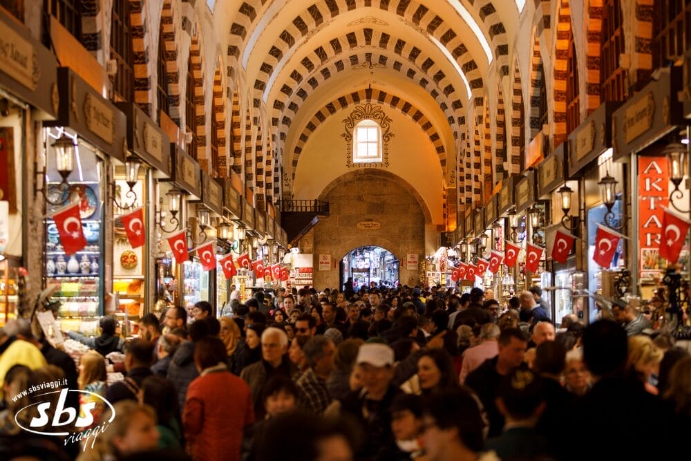 Un affollato mercato coperto con soffitti ad arco, fiancheggiato da negozi e bandiere turche. Molte persone camminano in questo spazio vivace, circondate da luci luminose, merci in vendita e dal ronzio delle macchine automatiche Bozza.