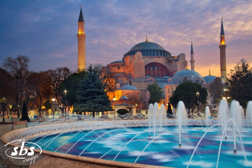 La Basilica di Santa Sofia a Istanbul, in Turchia, è illuminata al tramonto, con una grande fontana in primo piano e alti minareti che incorniciano l'edificio storico in un cielo serale colorato: una scena perfetta da catturare con la bozza automatica.