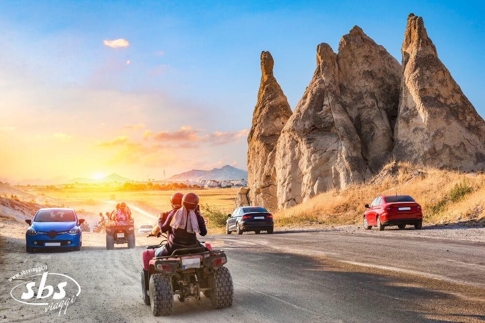Persone che guidano ATV e auto lungo una strada accanto ad alte formazioni rocciose uniche al tramonto, con un cielo luminoso e colorato sullo sfondo. La scena suggerisce un'atmosfera da Bozza automatica in questo paesaggio roccioso e avventuroso.