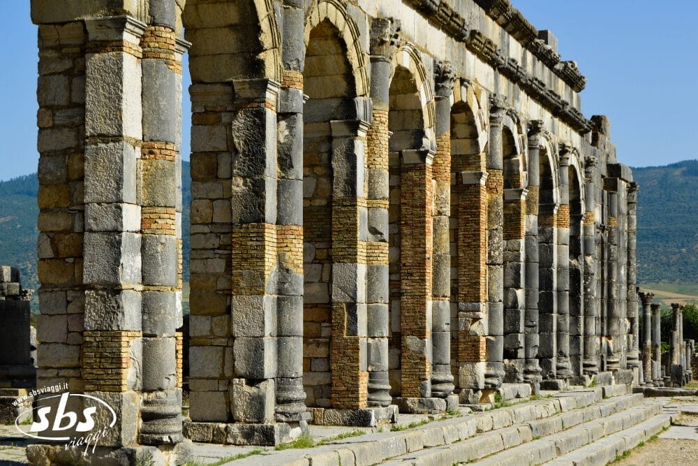 Sotto il cielo del Marocco, una fila di antichi archi in pietra si staglia alla luce del sole, con i loro blocchi consumati dalle intemperie che ricordano la grandezza delle Città Imperiali, con le montagne lontane che arricchiscono questo paesaggio storico. Perfetto per un'avventura Fly & Drive.
