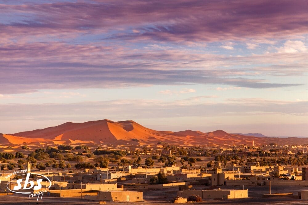 Una città del deserto con edifici dai tetti piatti si trova ai margini di grandi dune di sabbia arancione, sotto un cielo drammatico e parzialmente nuvoloso al tramonto. Il paesaggio è immerso in una luce calda e dorata, una vera e propria Bozza automatica dell'arte della natura.