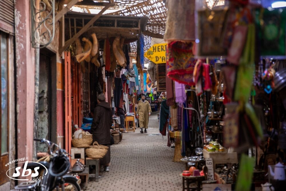 Una stretta e vivace strada del mercato del Marocco, fiancheggiata da tessuti colorati, oggetti appesi, cesti e oggetti di artigianato. Le persone camminano lungo il sentiero di ciottoli mentre la luce del sole filtra attraverso le grate di legno, evocando il fascino di un riad tradizionale.
