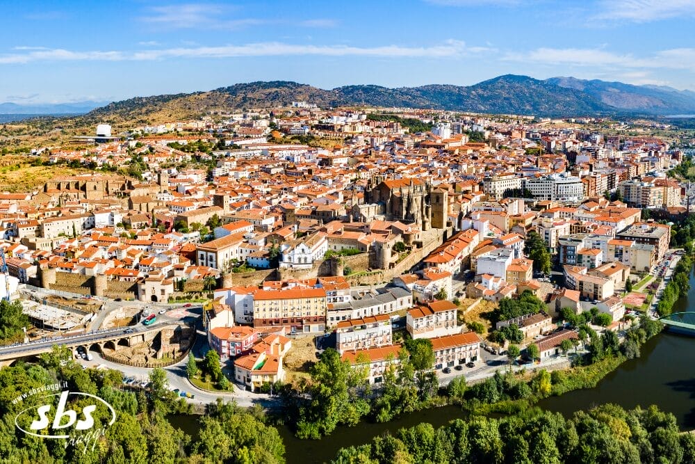 Veduta aerea di una città storica con edifici dai tetti rossi, strutture in pietra e un fiume in primo piano: la bozza automatica perfetta per immortalare colline verdi e montagne lontane sotto un cielo azzurro e limpido.