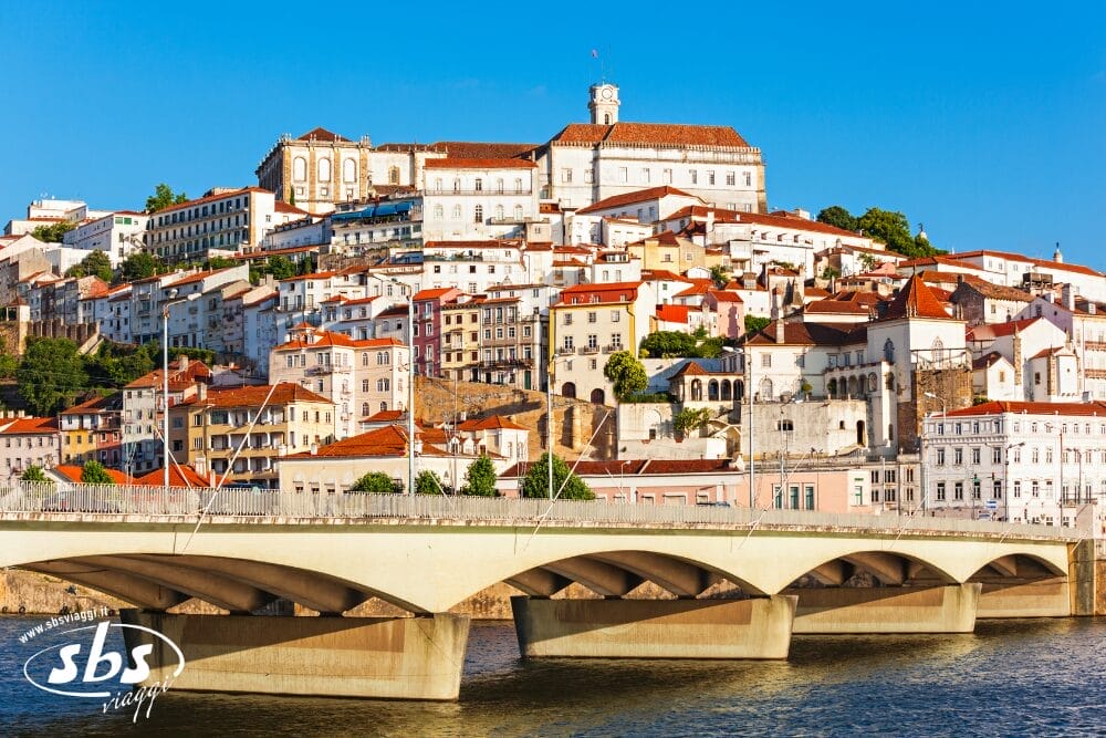 Una veduta di Coimbra, in Portogallo, che mostra edifici dai tetti bianchi e rossi accatastati su una collina, con la storica università in cima e un ponte che attraversa il fiume Mondego in primo piano sotto un cielo azzurro e limpido: una scena ideale per una Bozza automatica.
