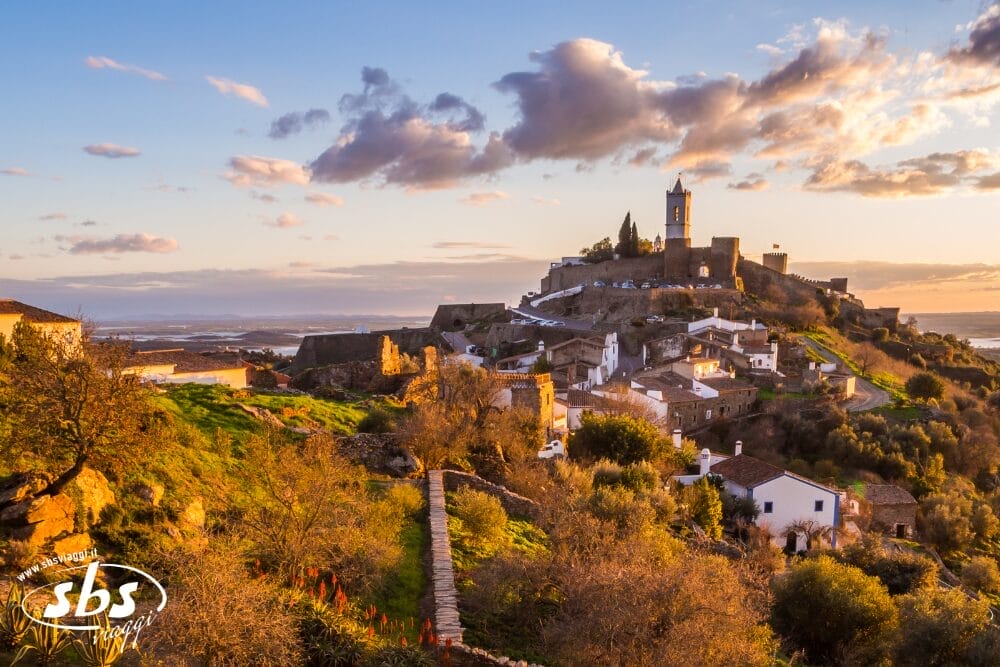 Borgo medievale in cima a una collina al tramonto con chiesa, muri in pietra e case bianche in mezzo a una vegetazione lussureggiante. La calda luce del sole bagna la scena, creando una Bozza automatica di edifici storici sotto un cielo parzialmente nuvoloso.