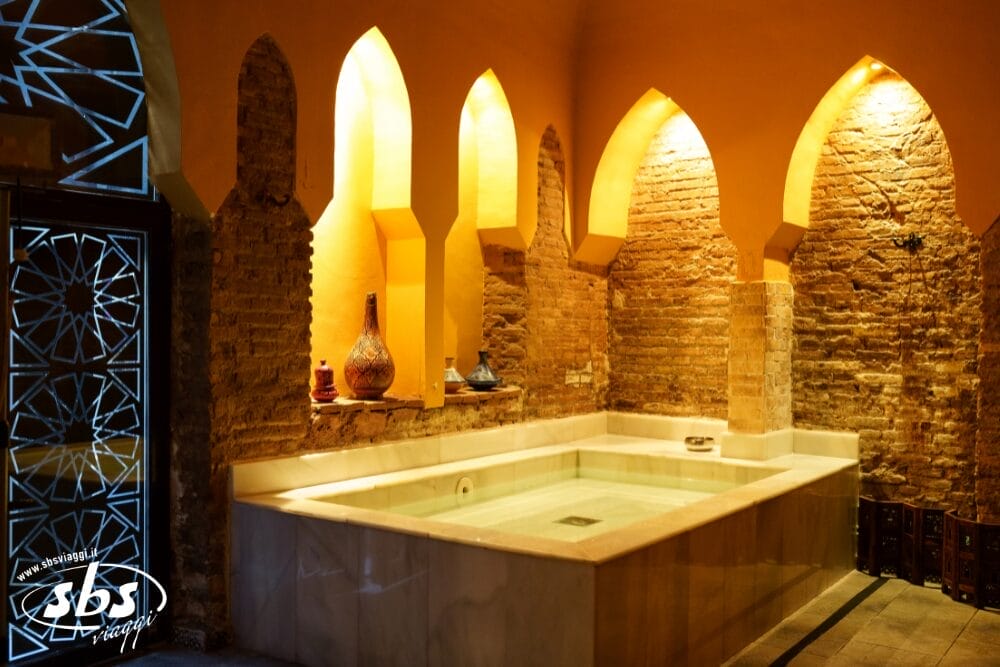 Un tradizionale hammam interno con una vasca a immersione decorata, pareti in mattoni a vista, nicchie ad arco con ceramiche, luci gialle soffuse e una porta di vetro decorativa sulla sinistra offre un ambiente rilassante di bozza automatica.
